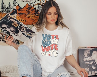 May include: A white t-shirt featuring a graphic design of the word "VOTE" repeated four times in a retro style with peace signs and red, white, and blue colours.