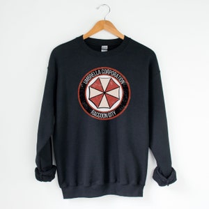 May include: Black sweatshirt with a distressed, circular logo featuring a red and white umbrella with the text "Umbrella Corporation Raccoon City".