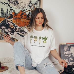May include: White t-shirt with a graphic of five different root vegetables and the text "Lets root for eachother".