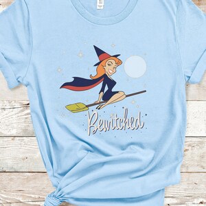 Op de afbeelding: Een lichtblauw T-shirt met een cartoonillustratie van een heks die op een bezem vliegt met de tekst "Bewitched" erop gedrukt.