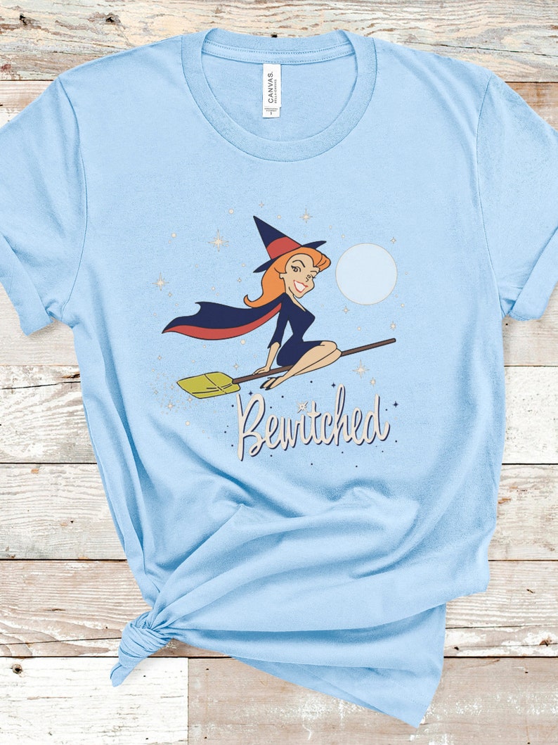Op de afbeelding: Een lichtblauw T-shirt met een cartoonillustratie van een heks die op een bezem vliegt met de tekst "Bewitched" erop gedrukt.