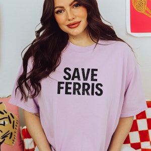 Pode incluir: Uma camiseta lavanda com o texto "SAVE FERRIS" impresso em preto.