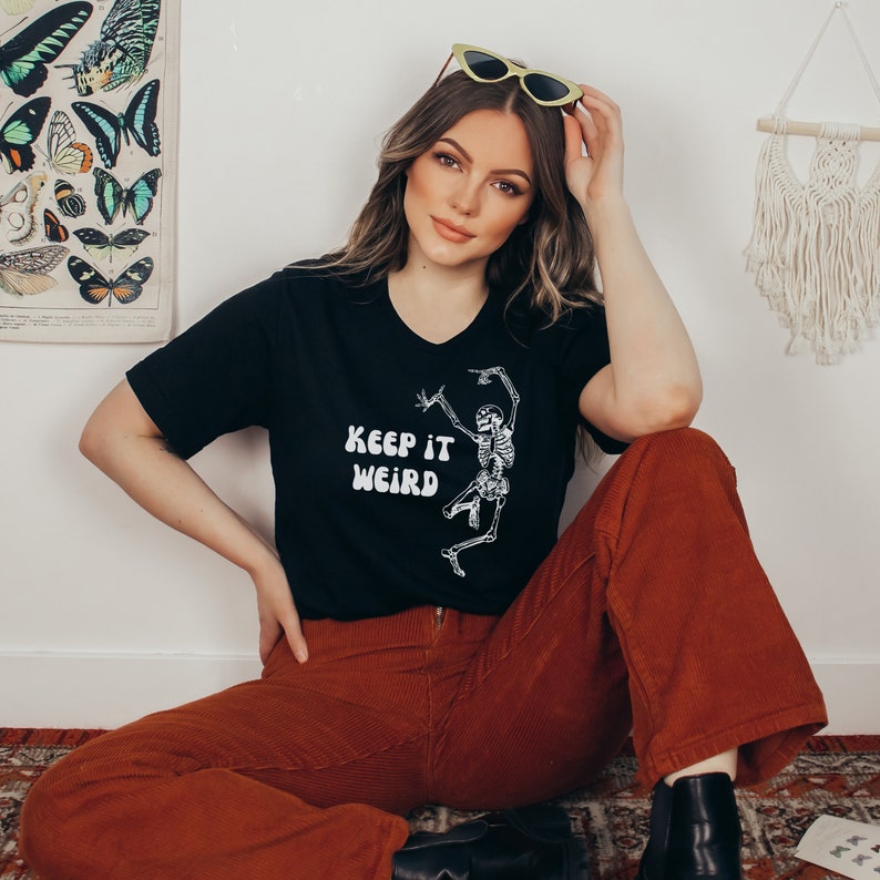 May include: A woman wearing a black t-shirt with a white skeleton graphic and the text "Keep it Weird". She is also wearing brown corduroy pants and black shoes.