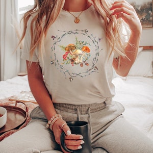 May include: A white t-shirt with a graphic of mushrooms and greenery in a wreath design. The shirt is being worn by a person sitting on a bed with a mug in their hand.