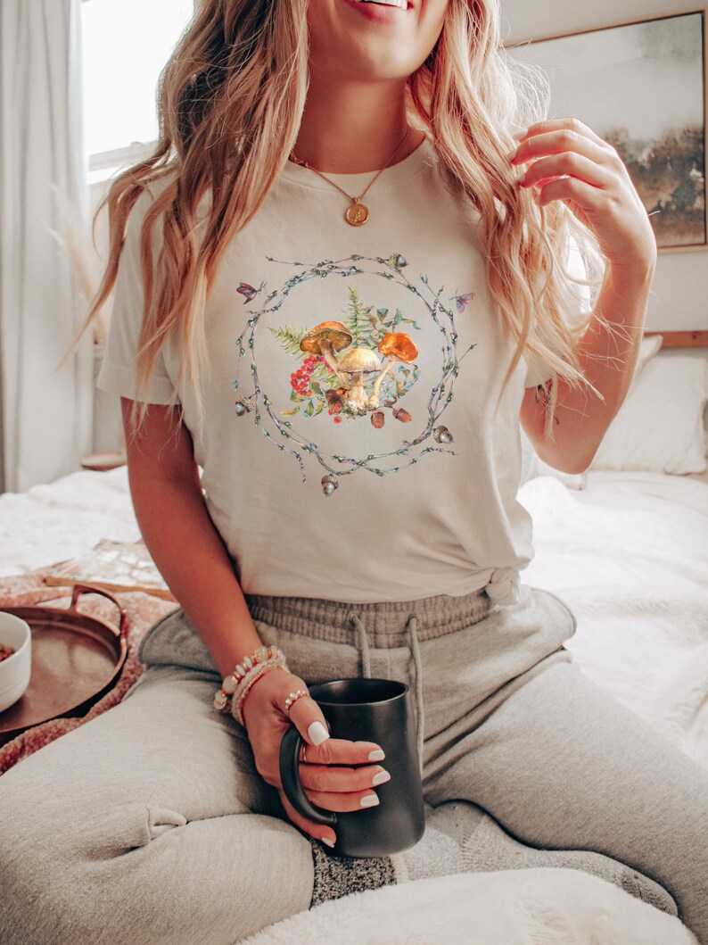 May include: A white t-shirt with a graphic of mushrooms and greenery in a wreath design. The shirt is being worn by a person sitting on a bed with a mug in their hand.