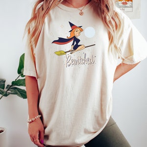 Op de afbeelding: Een cr&egrave;mekleurig T-shirt met een cartoonafbeelding van een heks die op een bezem vliegt. De heks draagt een zwarte jurk en een heksenhoed. De tekst "Bewitched" is onder de afbeelding gedrukt.