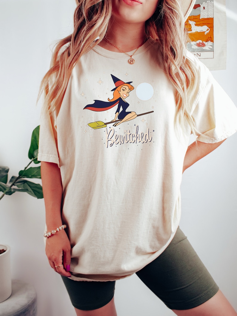 Op de afbeelding: Een cr&egrave;mekleurig T-shirt met een cartoonafbeelding van een heks die op een bezem vliegt. De heks draagt een zwarte jurk en een heksenhoed. De tekst "Bewitched" is onder de afbeelding gedrukt.