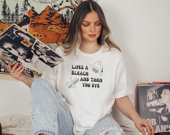 May include: A white t-shirt with the text "Lifes a bleach and then you dye" printed on it. The text is in a retro font and includes illustrations of a hairdryer, scissors, and a comb.