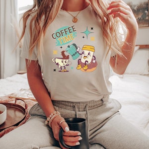 Op de afbeelding: Een wit T-shirt met een cartoonillustratie van een koffiekan die koffie in een koffiekopje giet. De tekst "COFFEE TIME" staat boven de illustratie. De persoon die het shirt draagt, houdt een zwarte mok vast.