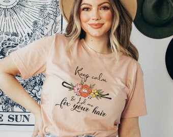 May include: A woman wearing a light pink t-shirt with the text "Keep calm & let me fix your hair" and a floral design featuring a hair clip.