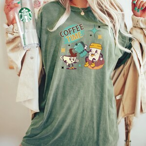 Op de afbeelding: Een oversized groen T-shirt met de tekst "COFFEE TIME" en een cartoonillustratie van een koffiekan die koffie in een koffiekopje giet. Het koffiekopje en de koffiekan hebben gezichten en armen.