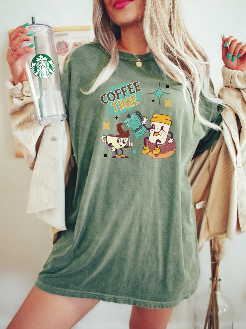 Op de afbeelding: Een oversized groen T-shirt met de tekst "COFFEE TIME" en een cartoonillustratie van een koffiekan die koffie in een koffiekopje giet. Het koffiekopje en de koffiekan hebben gezichten en armen.