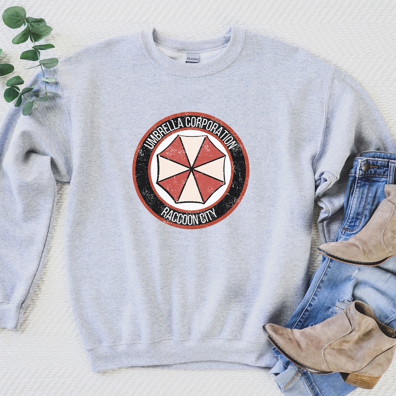 May include: A gray sweatshirt with a distressed, black and white logo featuring a red and white umbrella with the text "Umbrella Corporation Raccoon City".