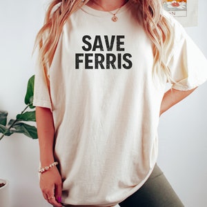 Pode incluir: Uma camiseta bege com o texto "SAVE FERRIS" impresso em preto. A camiseta &eacute; oversized e tem um caimento relaxado. A modelo est&aacute; usando a camiseta com shorts de ciclista pretos.