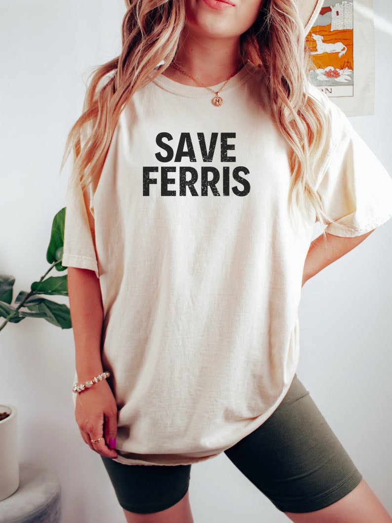 Pode incluir: Uma camiseta bege com o texto "SAVE FERRIS" impresso em preto. A camiseta &eacute; oversized e tem um caimento relaxado. A modelo est&aacute; usando a camiseta com shorts de ciclista pretos.