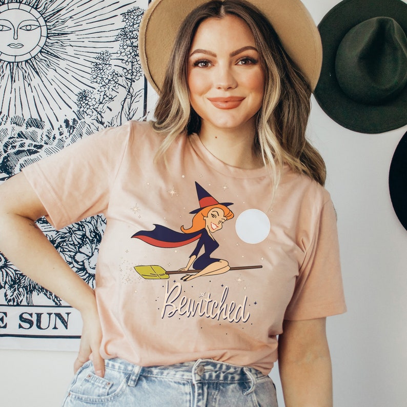 Op de afbeelding: Een vrouw draagt een lichtroze T-shirt met een afbeelding van een cartoonheks die op een bezem vliegt. De afbeelding bevat de tekst "Bewitched".