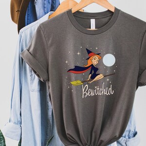 Op de afbeelding: Een donkergrijs T-shirt met een cartoonillustratie van een heks die op een bezem vliegt met een volle maan op de achtergrond. De tekst "Bewitched" is onder de heks gedrukt.