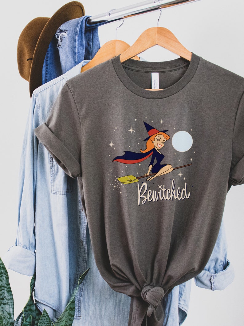 Op de afbeelding: Een donkergrijs T-shirt met een cartoonillustratie van een heks die op een bezem vliegt met een volle maan op de achtergrond. De tekst "Bewitched" is onder de heks gedrukt.