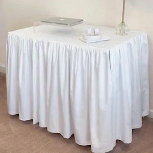 Può includere: Gonna da tavolo bianca con un bordo arricciato. La gonna è drappeggiata su un tavolo con un piano in vetro trasparente e un computer portatile sopra il tavolo.