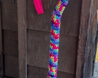 May include: A colorful crocheted cane with a pink handle and a pink crocheted wrist strap. The cane is leaning against a wooden wall.