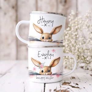 Può includere: Due tazze di smalto bianco con illustrazioni di conigli e nomi personalizzati. La tazza in alto dice "Henry -x-" e la tazza in basso dice "Everlyn -x-" con il testo "buona Pasqua" sotto il coniglio.