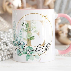 May include: White ceramic mug with a pink rim and handle. The mug features a gold circle with a gold letter "E" and green foliage. The text "Elizabeth" is written in cursive below the circle.