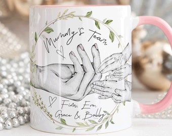 May include: White ceramic mug with a pink handle. The mug features a hand-drawn illustration of hands holding each other, surrounded by a wreath of green leaves. The text "Mommy's Team" is written in cursive with a heart symbol. Below the hands, the text "Finn, Eve, Grace & Bobby" is written in cursive.