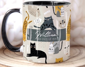 May include: A white ceramic mug with a black handle and black rim. The mug features a pattern of cats, milk cartons, and the words "Meow" and "Milk". The text "William aka crazy cat man" is printed on a gray band around the mug.