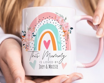 Può includere: Tazza in ceramica bianca con un design arcobaleno rosa, arancione e blu e dettagli floreali. Il testo "This Mummy is loved by Joey & Maisie" è stampato sulla tazza.