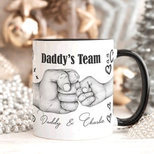 Puede incluir: Una taza de cerámica blanca con asa y borde negros, que presenta una ilustración en tinta negra de un puño adulto chocando con el puño de un niño. El texto "Daddy's Team" aparece arriba y "Daddy & Charlie" en letra cursiva debajo.