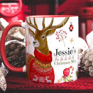 May include: White ceramic Christmas mug with a red rim and handle. The mug features a watercolour illustration of a reindeer wearing a red and white sweater, surrounded by Christmas-themed elements like berries, stars, and candy canes. The text "Jessie's Fa-la-la-la-la Christmas Mug" is printed on the mug.