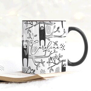 May include: White ceramic mug with a black handle. The mug features a black and white illustration of sloths hanging from branches with leaves and the name "Caroline" written in cursive.