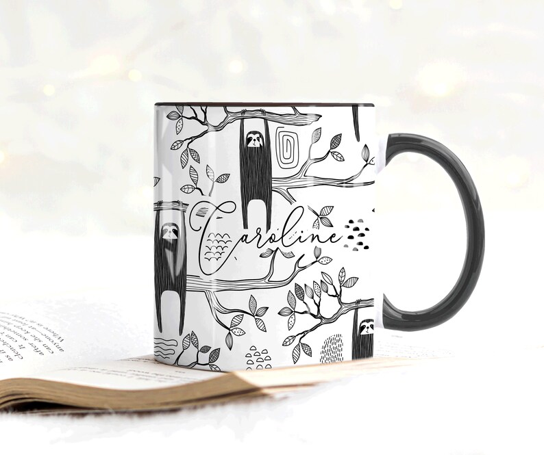 May include: White ceramic mug with a black handle. The mug features a black and white illustration of sloths hanging from branches with leaves and the name "Caroline" written in cursive.