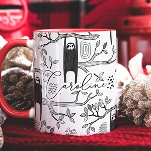 May include: White ceramic mug with a red handle. The mug has a black and white illustration of sloths hanging from branches with the name "Caroline" written in cursive.