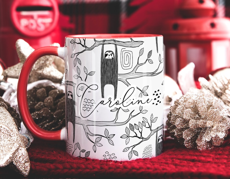 May include: White ceramic mug with a red handle. The mug has a black and white illustration of sloths hanging from branches with the name "Caroline" written in cursive.