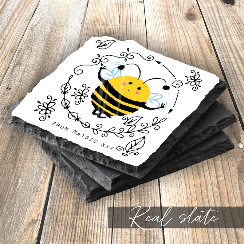 May include: A set of four black slate coasters with a white square tile on top. The tile features a cartoon bee with black and yellow stripes, surrounded by black floral designs. The text "From Maisie xxx" is written in black at the bottom of the tile.