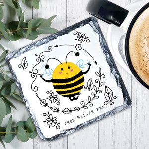 May include: A white slate coaster featuring a black and yellow cartoon bee illustration. The bee is surrounded by black floral designs and the text "FROM MAISIE XXX".