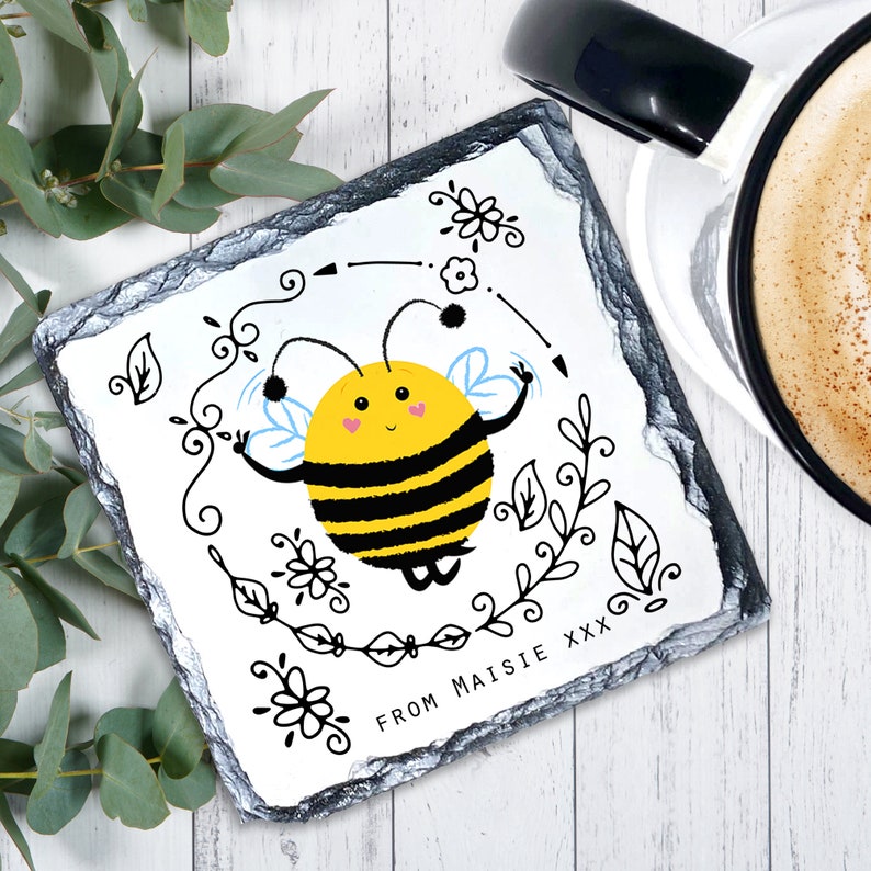 May include: A white slate coaster featuring a black and yellow cartoon bee illustration. The bee is surrounded by black floral designs and the text "FROM MAISIE XXX".