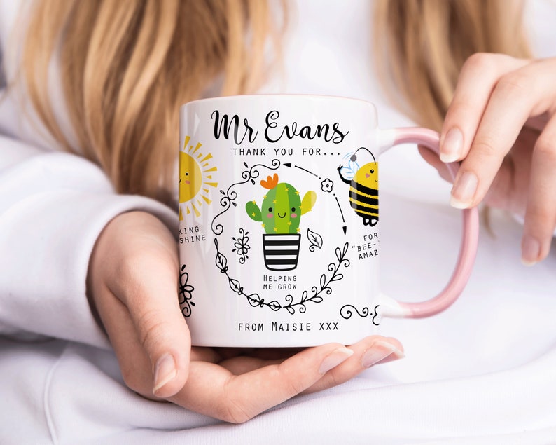 May include: White ceramic mug with a pink handle. The mug has a black and white illustration of a cactus with a smiling face, a sun, and a bee. The text on the mug reads "Mr Evans Thank you for... Helping me grow... For "bee-amaz" From Maisie XXX".