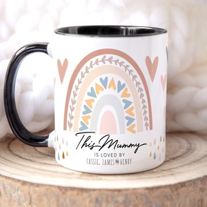 May include: White ceramic mug with a black handle. The mug features a rainbow design with hearts and leaves in shades of pink, orange, yellow, blue, and gray. The text "This Mummy is loved by Cassie, James & Henry" is printed on the mug.