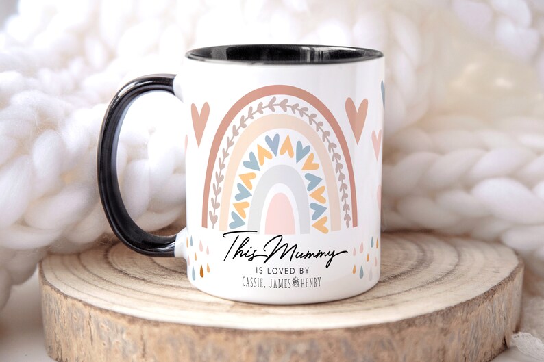 May include: White ceramic mug with a black handle. The mug features a rainbow design with hearts and leaves in shades of pink, orange, yellow, blue, and gray. The text "This Mummy is loved by Cassie, James & Henry" is printed on the mug.