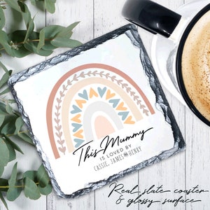 May include: A white square slate coaster with a rainbow design in pastel colors and a message that reads "This Mummy is loved by Cassie, James & Henry".