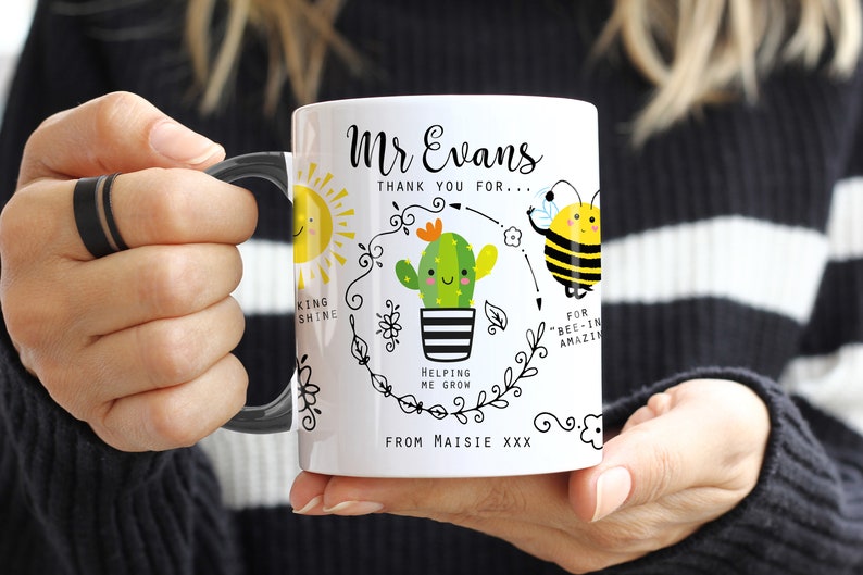 May include: White ceramic mug with a black handle. The mug features a black and white illustration of a sun, a cactus, a bee, and a flower. The text on the mug reads "Mr Evans Thank you for... King Shine Helping me grow For "Bee-in" Amazin From Maisie XXX"
