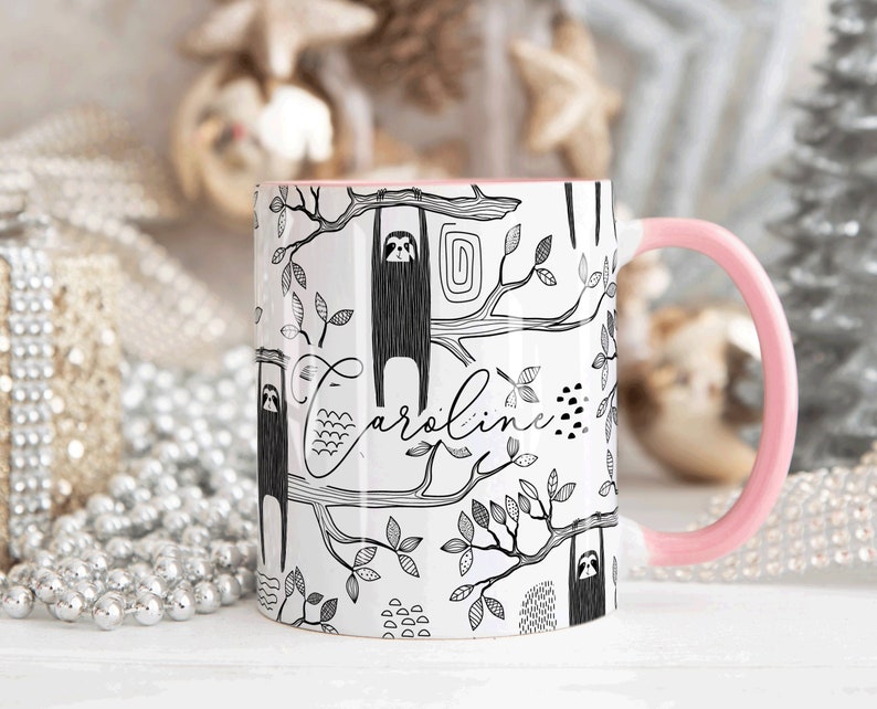 May include: White ceramic mug with a pink handle. The mug features a black and white illustration of sloths hanging from branches with the name "Caroline" written in cursive.