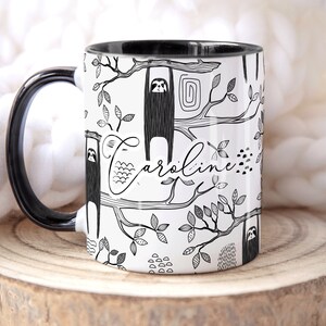 May include: White ceramic mug with a black handle and black rim. The mug features a black and white illustration of sloths hanging from branches with leaves. The name "Caroline" is written in cursive on the mug.