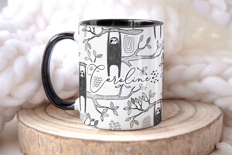 May include: White ceramic mug with a black handle and black rim. The mug features a black and white illustration of sloths hanging from branches with leaves. The name "Caroline" is written in cursive on the mug.