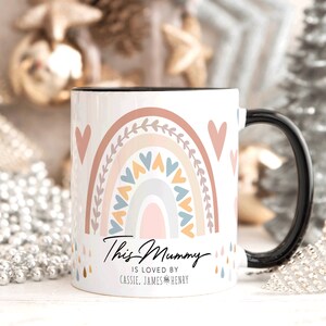May include: White ceramic mug with a black handle. The mug features a rainbow design with pink, orange, yellow, blue, and gray colors. The rainbow is surrounded by hearts and leaves. The text "This Mummy is loved by Cassie, James & Henry" is printed on the mug.