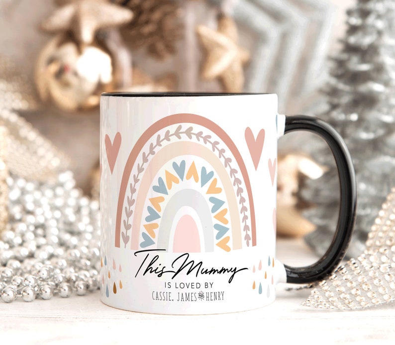 May include: White ceramic mug with a black handle. The mug features a rainbow design with pink, orange, yellow, blue, and gray colors. The rainbow is surrounded by hearts and leaves. The text "This Mummy is loved by Cassie, James & Henry" is printed on the mug.