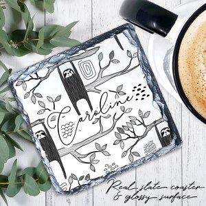 May include: Black and white illustrated coaster featuring sloths hanging from trees with the name "Caroline" written in cursive. The coaster is made of real slate with a glossy surface.