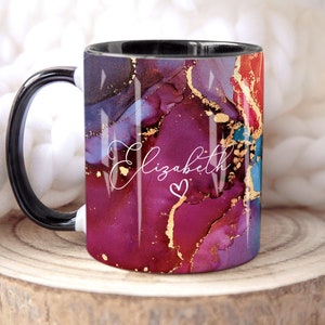 May include: A black and white mug with a black handle. The mug has a colorful abstract design with gold accents. The name "Elizabeth" is written in white script on the mug with a small heart.